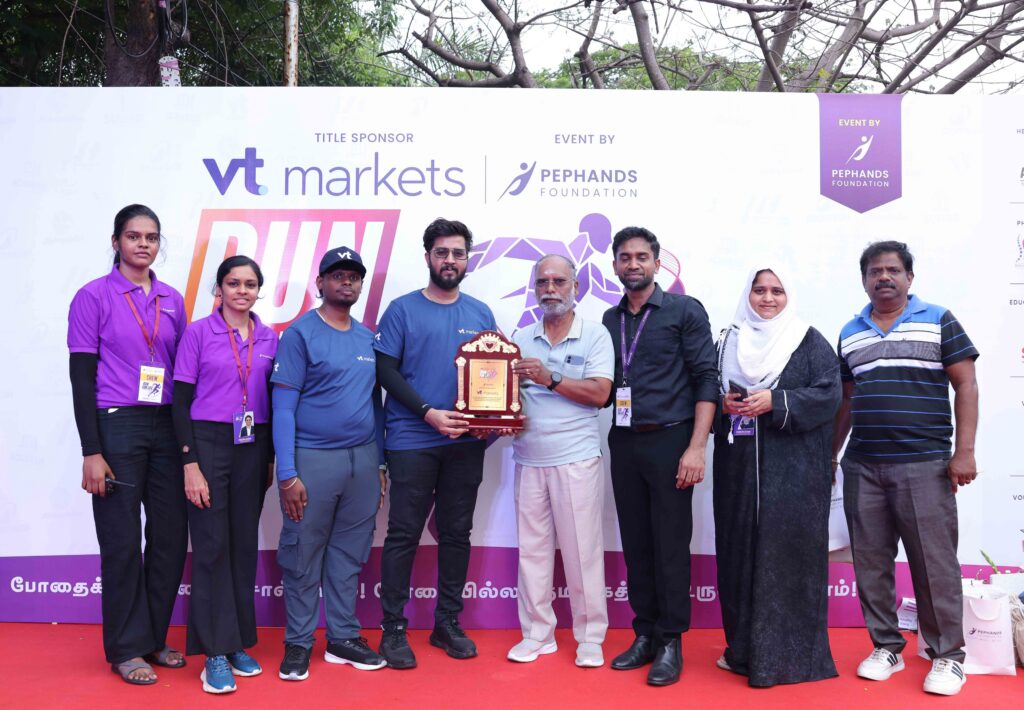 Group of people on a red stage posing with a trophy in front of a sponsorship banner at an event