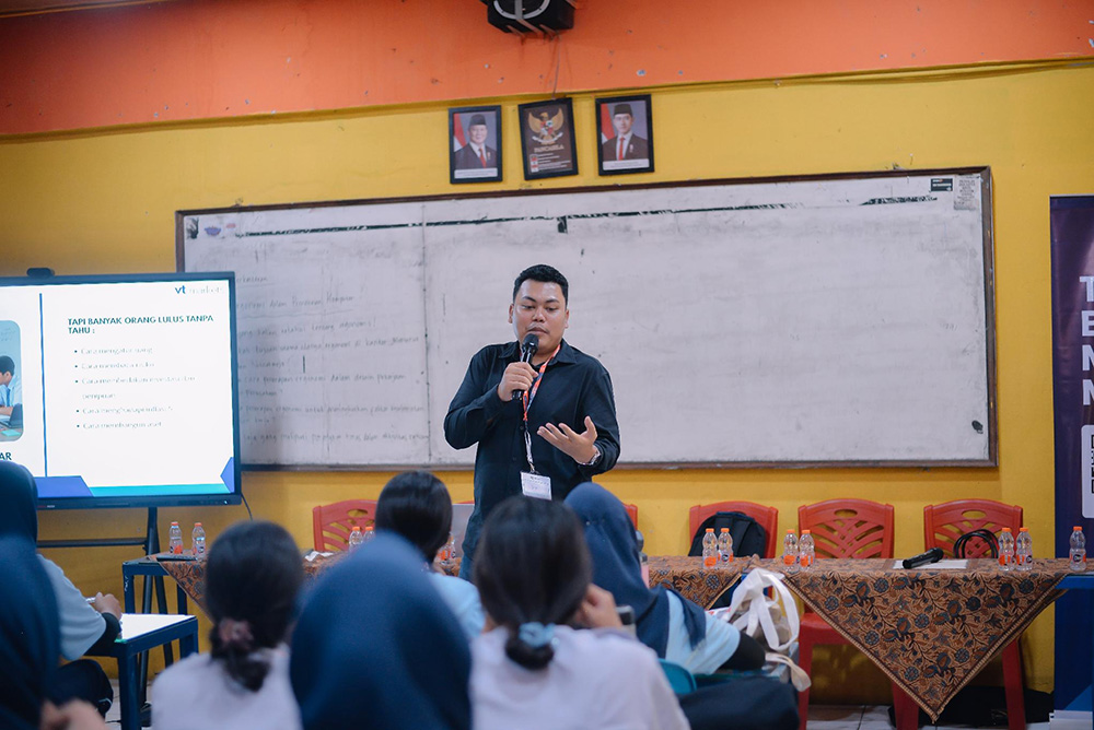 A man speaks into a handheld microphone at a classroom presentation addressing seated students with a slide on the screen to the left